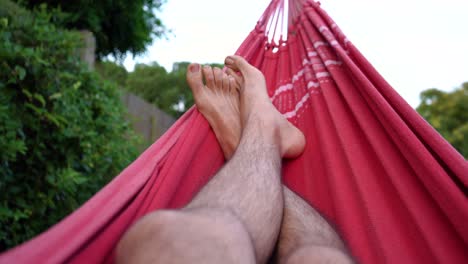 Füße-Und-Haarige-Beine-Eines-Mannes,-Der-Sich-Auf-Einer-Schwingenden-Roten-Hängematte-Im-Hinterhof-Entspannt