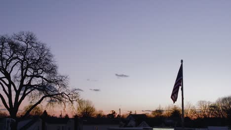 Eine-Luftaufnahme,-In-Geringer-Höhe-Der-Amerikanischen-Flagge,-Die-Während-Eines-Wunderschönen-Sonnenuntergangs-Im-Wind-Weht,-In-Zeitlupe-Mit-Der-Silhouette-Von-Bäumen-Im-Hintergrund