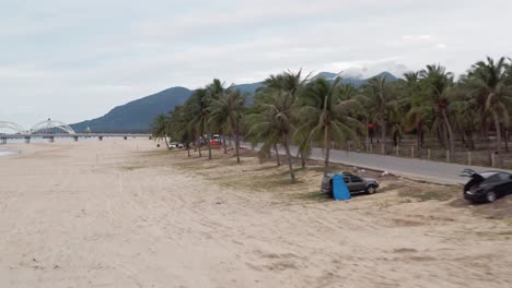 Camper-Vans-and-recreation-vehicles-parked-under-palm-trees-on-tropical-beach-side-road,-with-mountain-covered-in-clouds-in-background