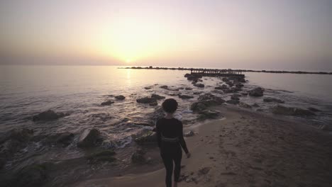 Schwarze-Afroamerikanische-Touristenfrau,-Die-Während-Des-Epischen-Sonnenuntergangs-über-Dem-Meer-Am-Felsigen-Strand-Von-Barcelona-Spanien,-Berühmtes-Touristenziel,-Spazieren-Geht