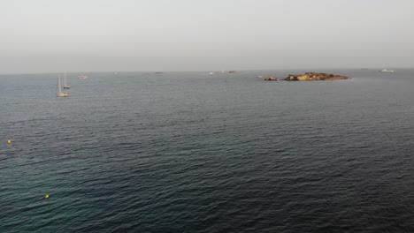 Entspannende-Luftaufnahme-Von-Zwei-Segelbooten-Und-Einer-Kleinen-Baleareninsel-Im-Mittelmeer-Mit-Ruhigen-Wellen-Am-Warmen-Sommernachmittagssonnenuntergang-In-Eivissa,-Ibiza,-Spanien