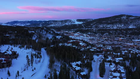 Aerial-Tilt-Down-Over-A-Quaint-Snow-Covered-Christmas-Town-Nestled-In-The-Mountains-With-Evergreen-Trees-And-pink-and-blue-skies