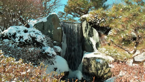 Statische-Aufnahme-Eines-Kleinen-Wasserfalls-Mit-Schneebedeckter-Umgebung-An-Sonnigen-Tagen