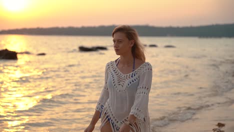 Beautiful-young-European-woman-in-blue-bikini-and-white-shirt-walks-in-the-surf-during-sunrise-on-a-sandy-beach-in-Thailand-while-looking-around
