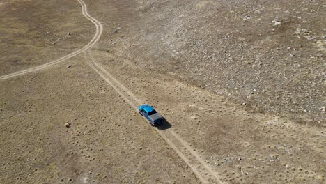 Blue-truck-driving-along-a-dirt-road-on-an-arid-desert-landscape---aerial-follow
