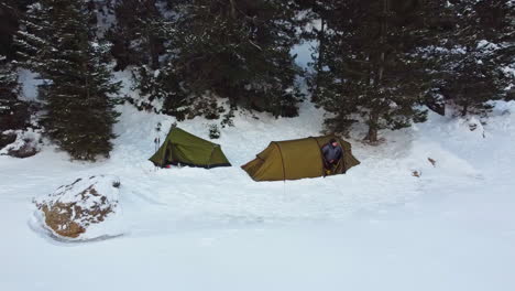 Pull-back-aerial-shot-from-a-camping-tents,over-a-frozen-lake,-covered-of-snow,at-the-background-a-mountain-full-of-snow,-revealing-forest,-while-men-getting-the-heads-out-of-tents-to-see-the-scenery
