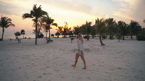 Young-blond-woman-in-summer-clothes-walks-barefoot-on-the-white-beach-among-the-green-palm-trees-during-a-beautiful-sunset