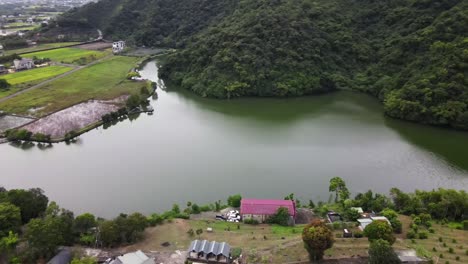 Green-watered-lake-at-base-of-lush-green-mountain-near-rural-housing-and-rice-paddy