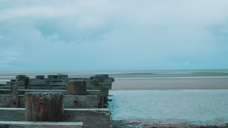 A-small,-wooden-structure-stretches-past-a-small-veins-of-water-and-sits-on-the-slowly-drying-beach-at-lowtide-whilst-on-the-horizon-an-angry-storm-begins-to-brew