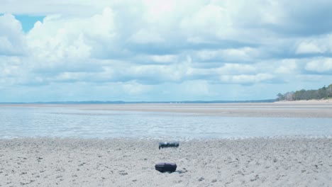 A-small-DJI-Mavic-Air-drone-takes-off-from-its-small-carry-case-and-flies-above-the-wet-sand-and-large-veins-of-water-stretching-along-the-low-tide-beach-and-into-the-large,-fluffy,-white-clouds