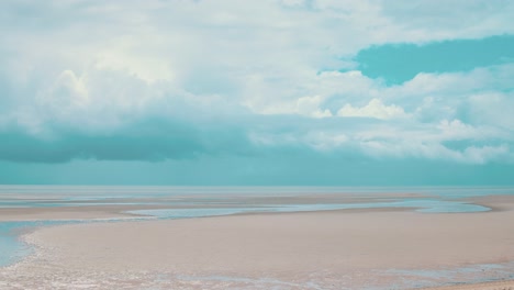 Timelapse-of-large-storm-clouds-brewing-on-the-horizon-of-a-beach-at-lowtide-where-large-veins-of-water-stretch-along-the-pale-sand-whilst-the-warm-sun-still-beams