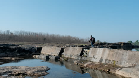 Unrecognizable-man-with-backpack-crossing-river-over-bridge-rubble