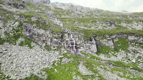 Kleiner-Wasserfall-In-Einer-Bergkette-Zwischen-Felsigen-Und-Grasbewachsenen-Steilen-Böden
