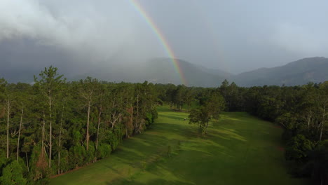Golf-course-aerial-view-with-a-beautiful-rainbow,-mountains-and-rainstorm