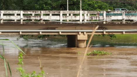 Flowing-muddy-river-under-a-bridge-with-assorted-traffic-and-tuktuk
