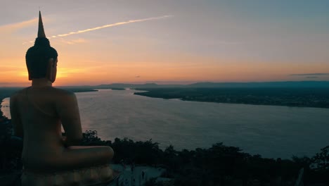 Imágenes-De-4k-De-Una-Impresionante-Puesta-De-Sol-Sobre-El-Poderoso-Río-Mekong-En-Laos,-Observada-Pacíficamente-Por-Una-Gran-Estatua-De-Buda