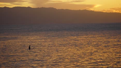 A-lone-surfer-sits-and-watches-the-sets-of-waves-from-the-Pacific-on-North-Shore-Oahu-just-after-sunset-with-orange-and-yellow-reflections-on-the-water