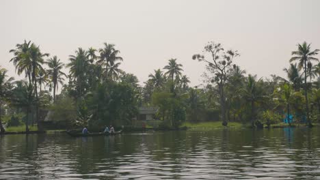 Die-Kamera-Segelt-An-Drei-Menschen-In-Der-Ferne-In-Einem-Kleinen-Kanalboot-Auf-Den-Backwaters-Von-Kerala-Mit-Palmen-Am-Ufer-Und-Der-Mittagssonne
