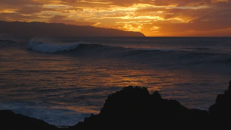 A-large-wave-rolls-in-as-the-sun-has-just-set-over-Hawaii's-north-shore-with-volcanic-rocks-on-the-beach-silhouetted-against-the-orange-light