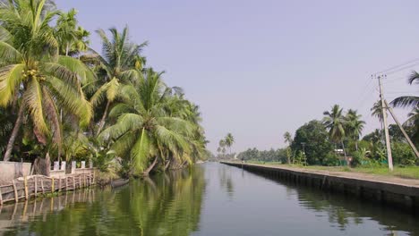 In-Der-Hitze-Des-Tages-Mit-Palmen-Bewegen-Sie-Sich-Langsam-Die-Kerala-Rückstaukanäle-Hinunter,-Mit-Traditionellen-Zäunen-Auf-Der-Einen-Seite-Und-Einem-Modernen-Betonweg-Auf-Der-Anderen-Seite