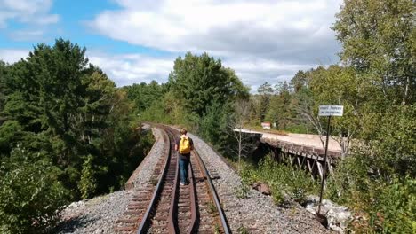Dolly-in-drone-shot-of-a-man-with-a-yellow-backpack-on-walking-on-a-train-rail-surrounded-by-pine-trees-in-Canada