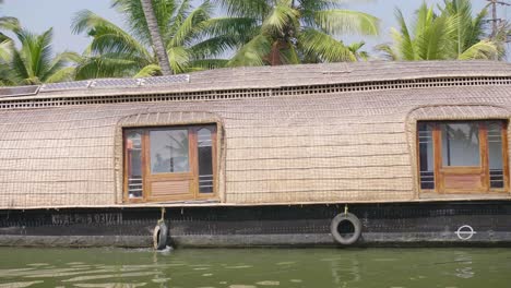 Ein-Elegantes-Und-Traditionelles-Hausboot-Schwimmt-In-Der-Hitze-Des-Tages-Auf-Dem-Kulturerbe-Der-Backwaters-Von-Kerala-Mit-Palmen-Im-Hintergrund-Vorbei