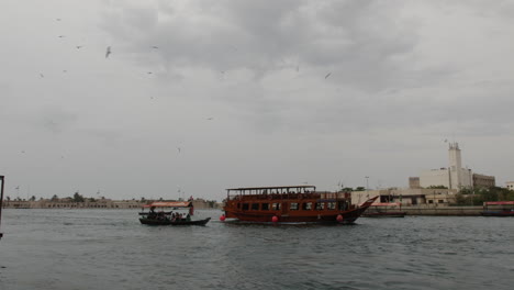 Barcos-De-Oriente-Medio-Bajando-Por-El-Río-En-El-Viejo-Dubai-En-Un-Día-Nublado-Y-Nublado-Mientras-Las-Gaviotas-Vuelan-Por-Encima-En-Cámara-Lenta-60fps