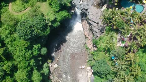 Bird's-eye-view-top-down-reveal-drone-shot-of-tegenungan-waterfall-and-a-river-surrounded-by-a-lush-green-jungle-and-resorts-in-Bali,-Indonesia