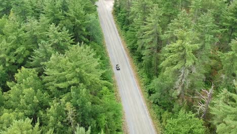 Downwards-tracking-drone-shot-of-someone-driving-an-ATV-or-four-wheeler-on-a-dirt-road-in-the-middle-of-the-forest-in-Canada