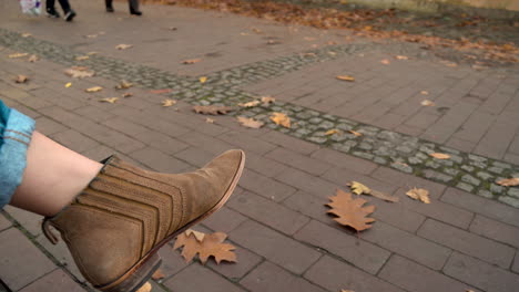 Woman's-Foot-In-Brown-Ankle-Boots-While-Sitting-With-People-Passing-By-At-Oliwski-Park-In-Gdansk,-Poland