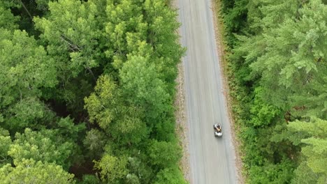 Drone-tracking-shot-going-upwards-of-someone-driving-an-ATV-on-a-dirt-road-surrounded-by-green-pine-trees-in-Canada