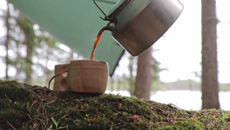 Coffee-poured-to-a-wooden-cup-from-a-steel-coffee-pot-in-a-camping-setup-in-Repovesi-national-park-in-Finland,-stationary-shot
