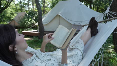 Girl-with-dark-hair-in-white-country-dress-reads-a-book-in-a-hammock