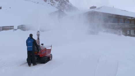 Person-clearing-a-trail-to-a-house-with-a-snow-blower-on-a-cloudy-day-in-the-alps