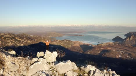 AERIAL---Person-hiking-raises-arms-in-mountains,-Lake-Skadar,-Montenegro,-reverse