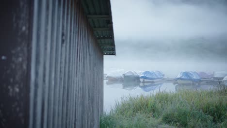 Mysterious-cabin-by-lakeside,-boats-moored-on-foggy-misty-lake