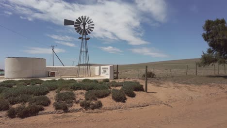 A-farm-wind-pump-slowly-rotates-in-a-light-breeze-in-arid-farmland,-slow-pan-to-the-right