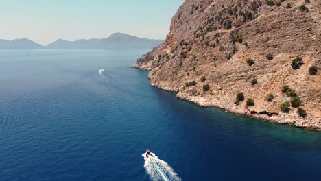 Aerial-dolly-shot-following-small-boat-in-beautiful-blue-waters-close-to-a-rocky-mountain
