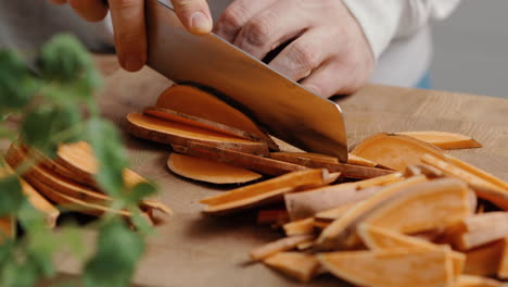 Male-chef-sliceing-sweet-potato-in-thin-slices-in-the-kitchen-on-a-wooden-cutting-board-in-the-kitchen