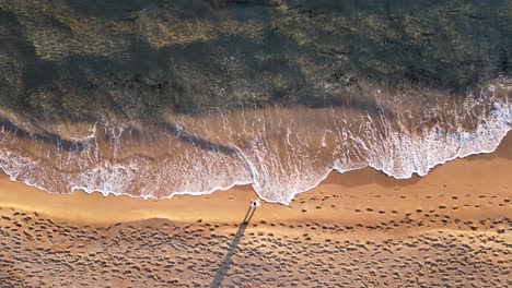 Top-down-aerial-view-of-waves-crushing-on-an-empty-beach-with-one-man-standing-in-the-middle