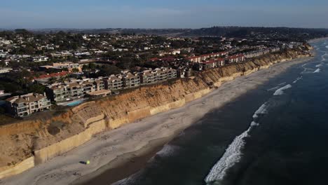 A-stunning-aerial-drone-shot,-flying-along-the-coastline-over-the-ocean-while-filming-the-beautiful-waves-hitting-the-shore,-Solana-Beach---San-Diego---California
