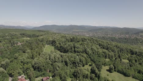Ascending-aerial-view-over-dense-forest-landscape-with-houses-far-in-the-distance