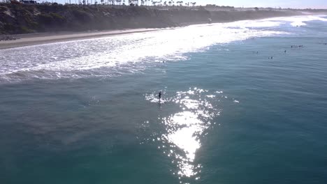 A-stunning-aerial-drone-shot-of-a-man-supping-in-the-ocean-close-to-the-beach,-Carlsbad-State-Beach---California