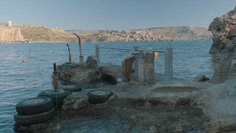 Meerwasser-Umgibt-Einen-Alten,-Verlassenen-Bootssteg-Im-Vordergrund-Mit-Den-Felsigen-Klippen-Einer-Maltesischen-Insel-Im-Hintergrund