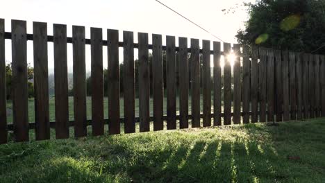 Sun-shining-behind-a-picket-fence-over-green-lawn-as-camera-pans-across