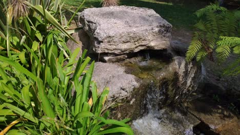 Fresh-water-gushing-from-the-fountain-among-granite-rocks,-ferns-and-decorative-plants-next-to-you-in-the-landscaped-park-on-a-sunny-summer-morning