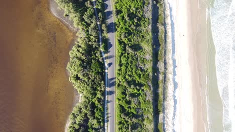 Drone-aerial-top-view-of-a-car-going-along-an-empty-road-between-the-beach-and-a-lake-in-Australia