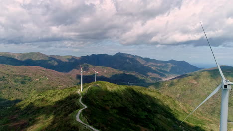 Aerial-view-of-wind-turbines-on-path-between-green-mountains-during-cloudy-day---Production-of-renewable-energy-in-nature