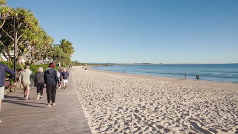 Beachside-Boardwalk-With-Tourists-Walking-Under-Pandanus-Trees-With-Ocean-View,-4K-Wide-Slow-Motion
