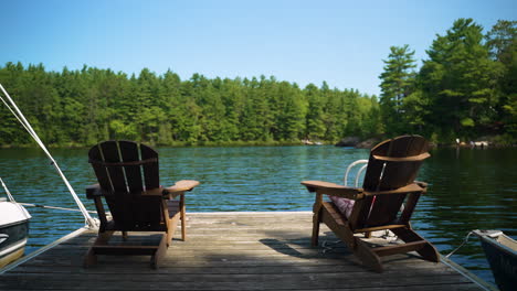 Two-empty-Muskoka-chairs-sit-at-the-end-of-a-dock-looking-out-at-the-water-on-a-calm,-beautiful-summer-day